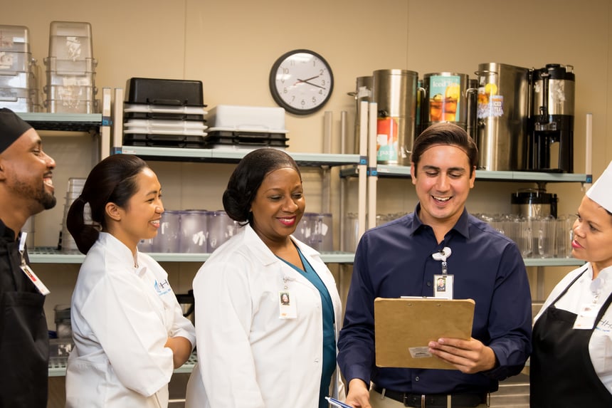 A manager holding a clipboard leads a team huddle with culinary staff in a hospital kitchen, all smiling and engaged in conversation.