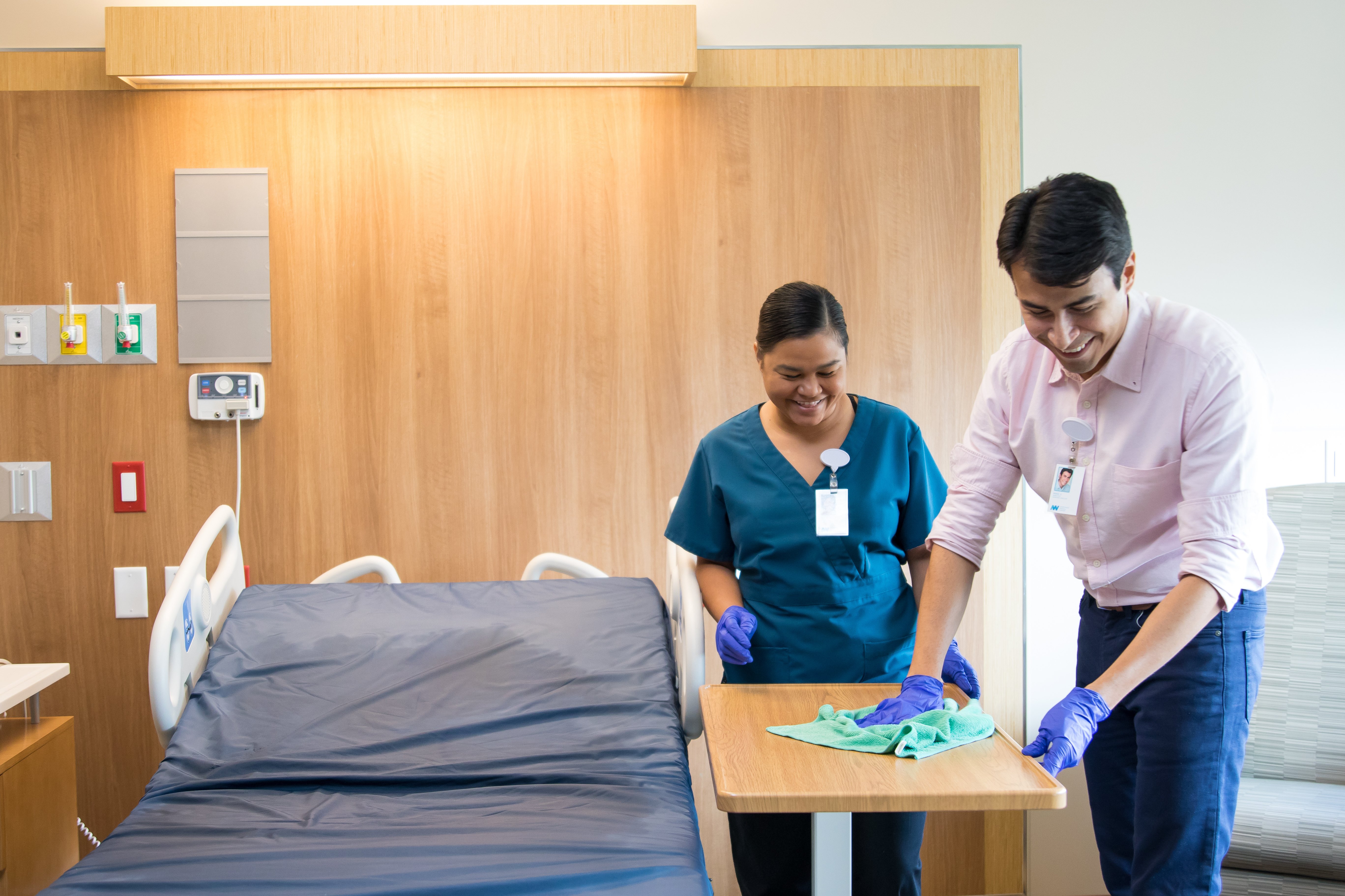 A healthcare manager in a pink shirt and blue gloves wipes down a bedside table alongside a team member in scrubs, both smiling during a hospital room cleaning.