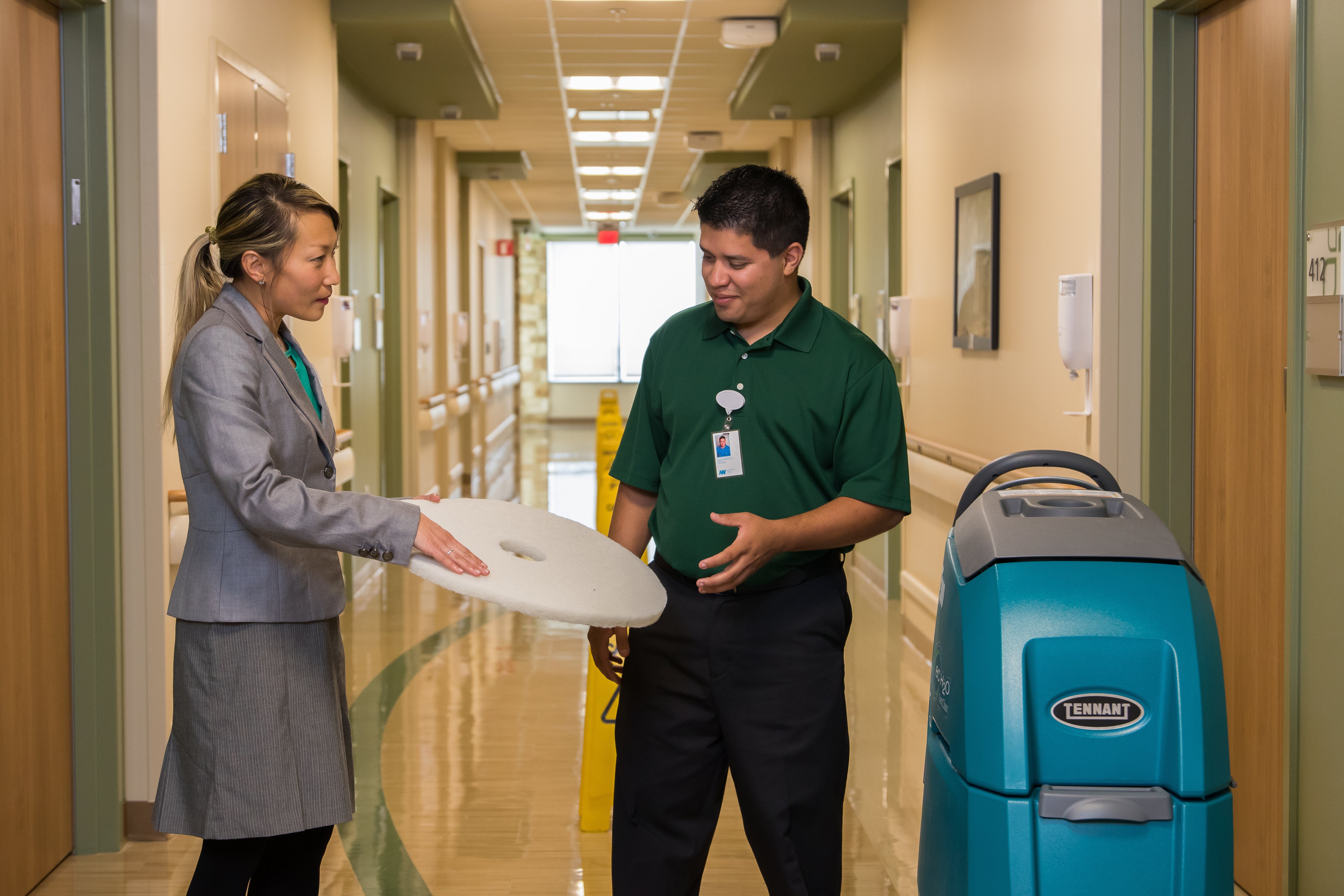 A manager in a gray blazer holds a floor buffer pad while speaking with a team member in a green polo shirt next to a floor cleaning machine in a hospital hallway.