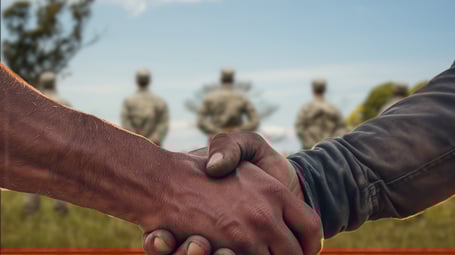 Gemini said A close-up photograph of a firm handshake between two individuals, with a group of blurred military personnel in uniforms standing in a field in the background.
