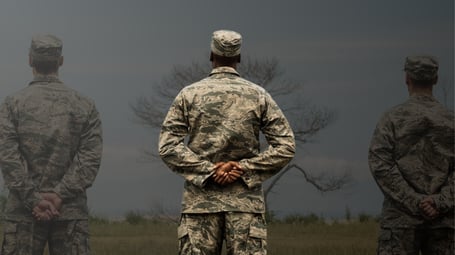 A composite photograph showing the rear view of three military personnel in, standing at attention in a field under a dark, moody sky with a bare tree. The central figure is in focus, while the two flanking figures are transparent, giving a ghost-like appearance.
