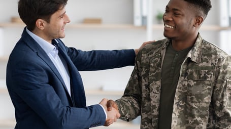 A professional photograph shows a smiling man in a blue suit and tie shaking hands with a smiling man in a camouflage military uniform. They are in an office setting. The man in the suit is on the left, and the man in the uniform is on the right, with his left hand on the other man's shoulder. The background is slightly blurred but shows bookshelves and a window.