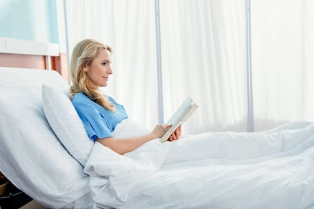 A blonde woman in a blue hospital gown sits propped up in a bed with white linens, holding an open book in her hands and gazing to the side.