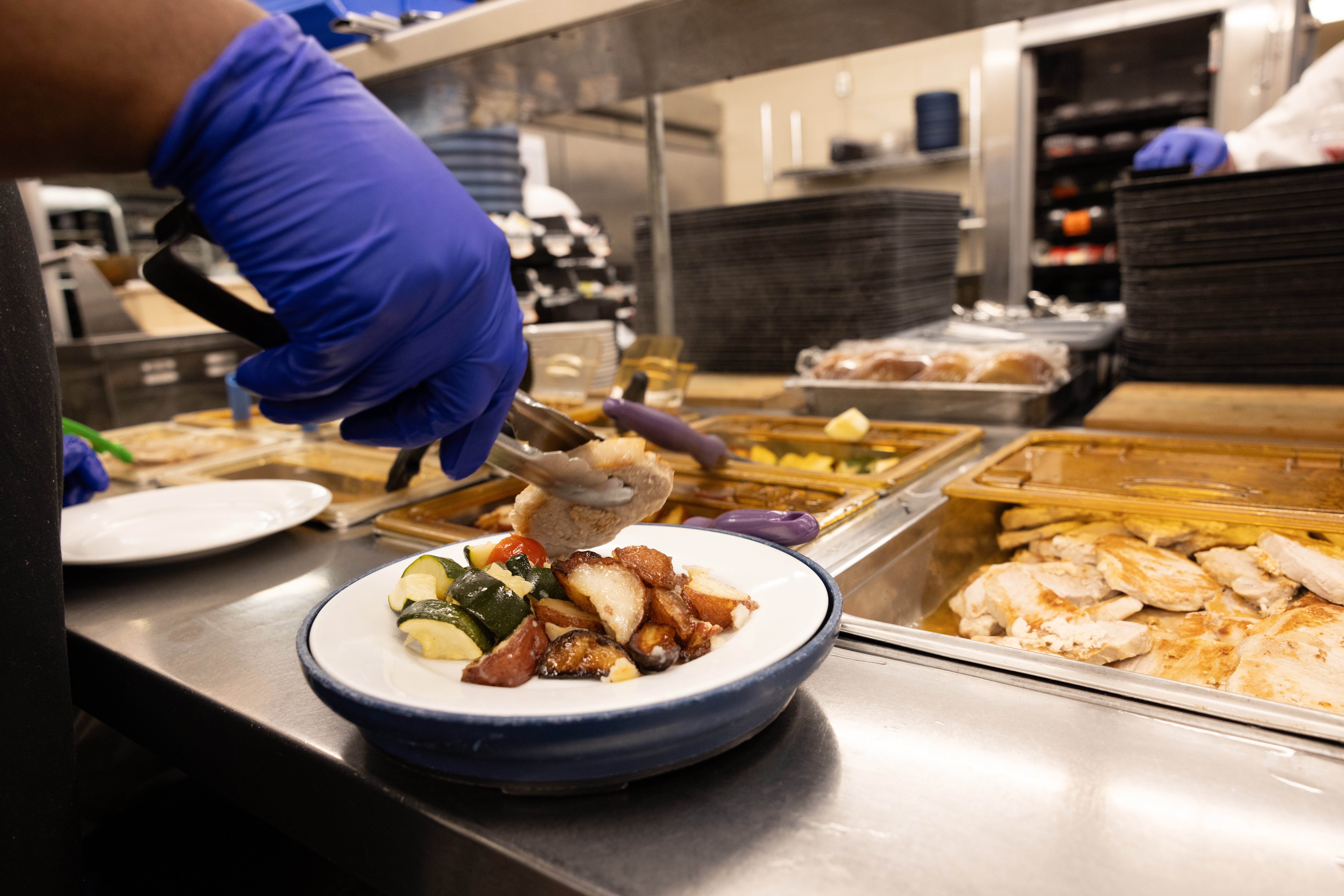HHS culinary team member plating roasted vegetables and pork in a hospital kitchen during patient meal preparation.