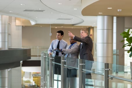 Three men in business attire stand on a glass balcony in a modern building, with one man in a dark suit pointing into the distance while another holds a tablet.