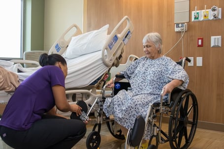 A female healthcare worker in a purple uniform kneels to adjust the footrests of a wheelchair for an elderly woman in a hospital gown.