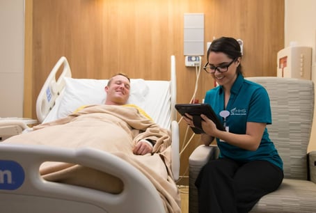 A smiling female HHS healthcare worker in a teal shirt uses a tablet while sitting beside a smiling male patient lying in a hospital bed covered with a beige blanket.