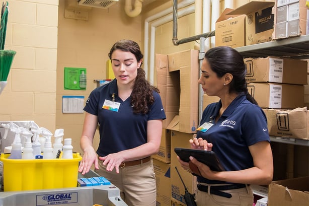 Two women in navy polo shirts stand in a storage room filled with cardboard boxes and cleaning supplies. One speaks, while the other holds a tablet, listening attentively.