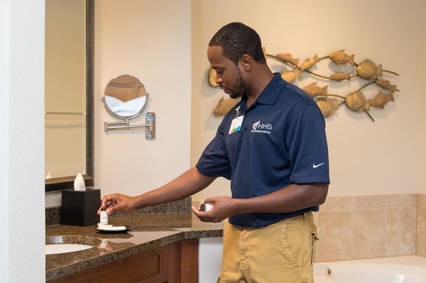 Man in a blue uniform cleaning a bathroom sink, holding a hotel soaps. A circular wall mirror and decorative fish art are visible in the background.