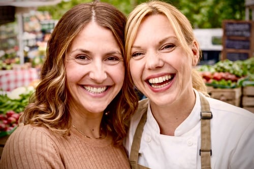 Two smiling women in front of a produce stand, showcasing an array of fresh fruits and vegetables.