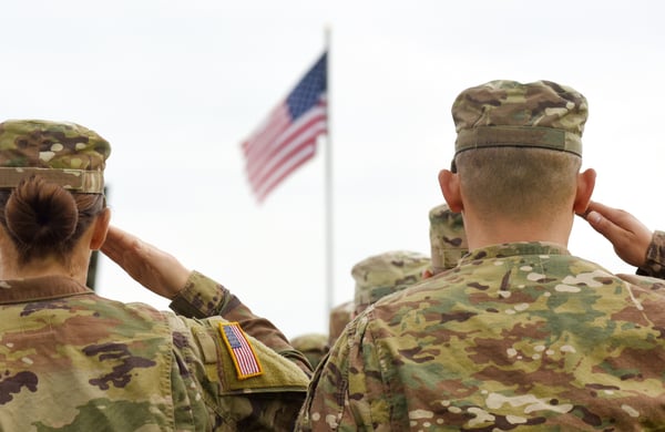 U.S. Army soldiers in camouflage uniforms stand in formation, saluting towards an American flag in the distance. The image is taken from behind, showing the backs of the soldiers, with one female soldier visible on the left. A U.S. flag patch is prominently displayed on the shoulder of one soldier. The setting is outdoors under an overcast sky, emphasizing the solemn and respectful nature of the scene.